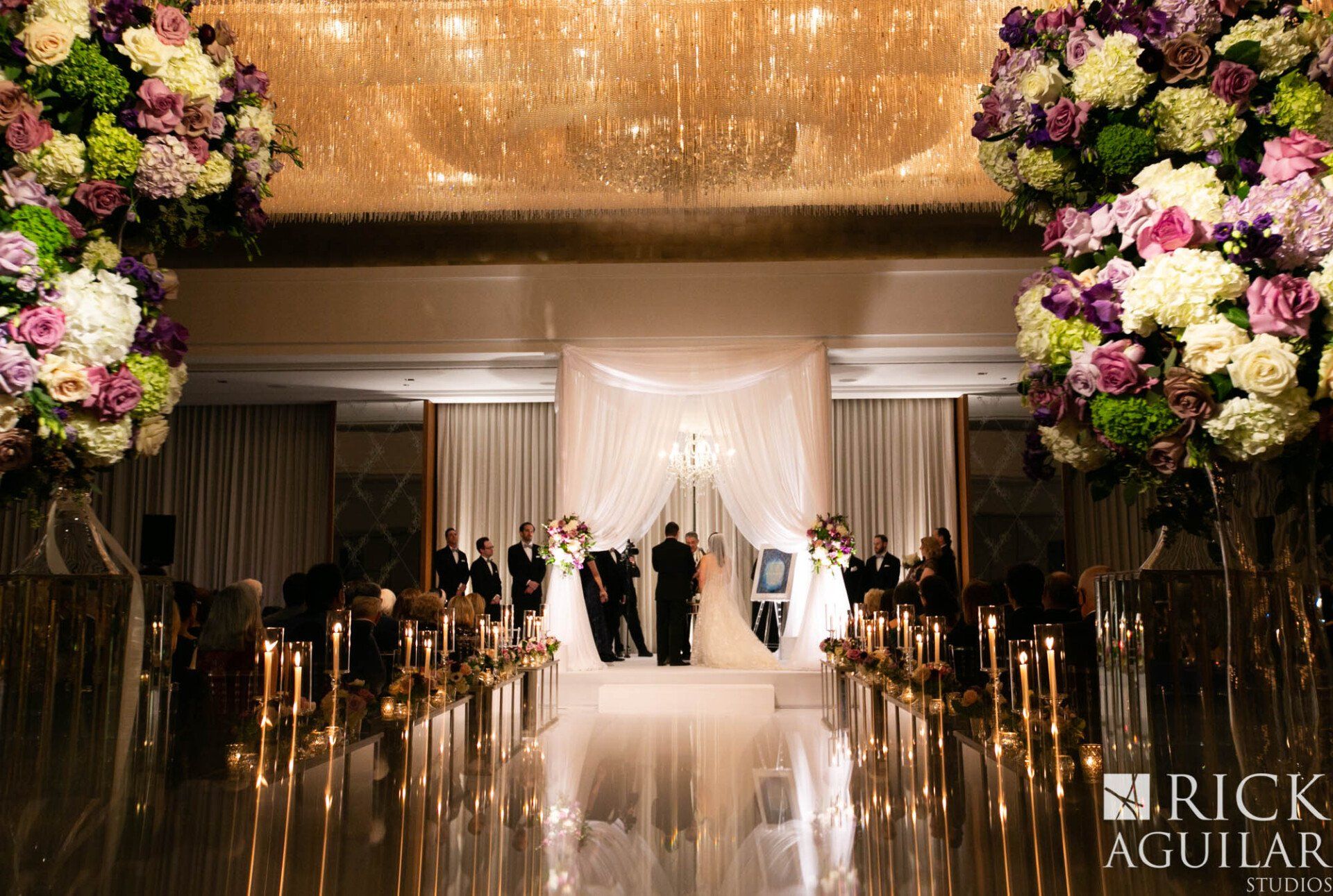 A wedding ceremony is taking place in a large room with flowers and candles.