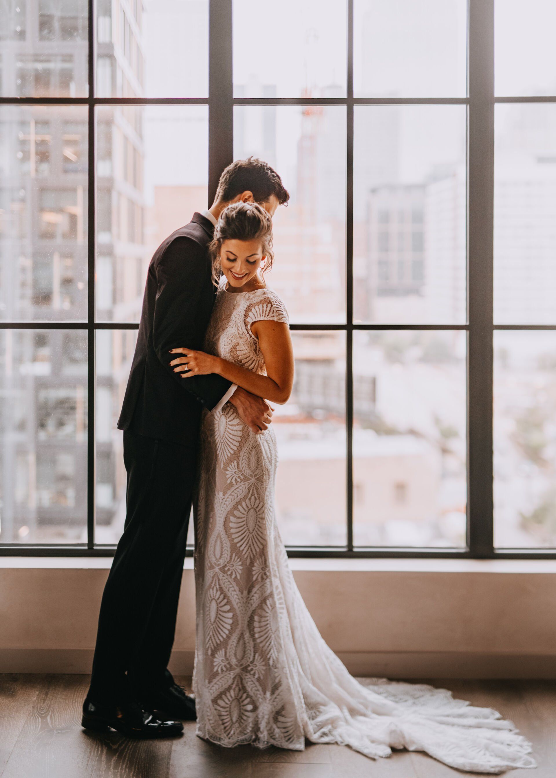 A bride and groom are hugging in front of a window.