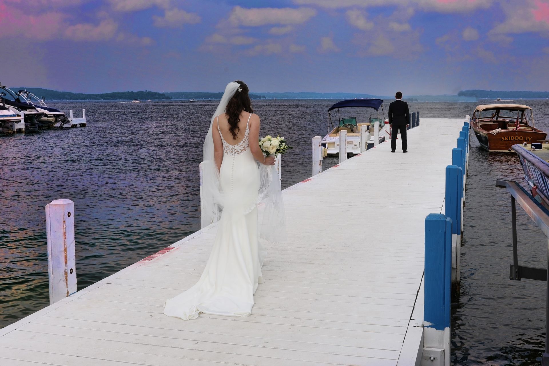 A bride and groom are standing on a dock with boats in the background.