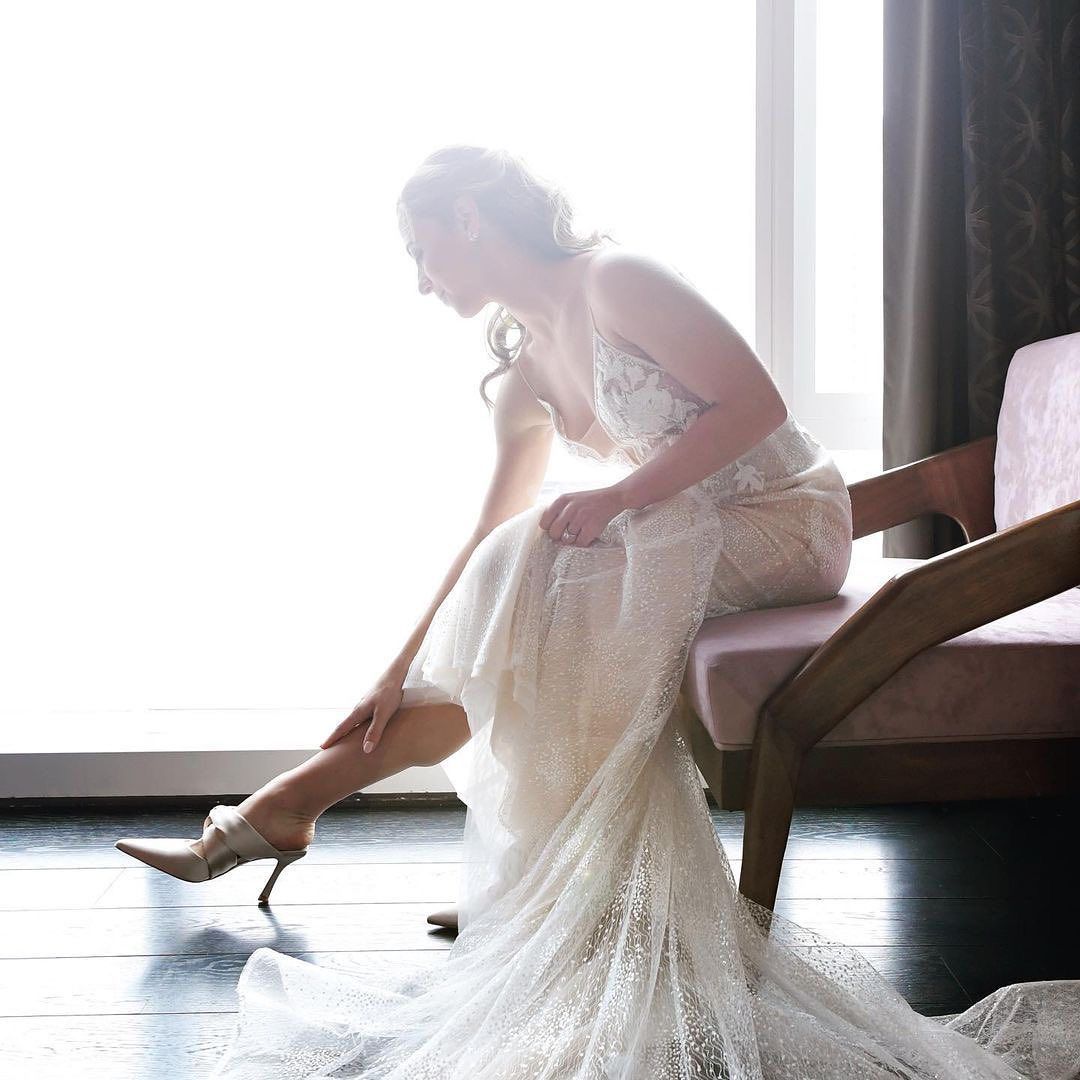 A woman in a wedding dress is sitting on a chair in front of a window.