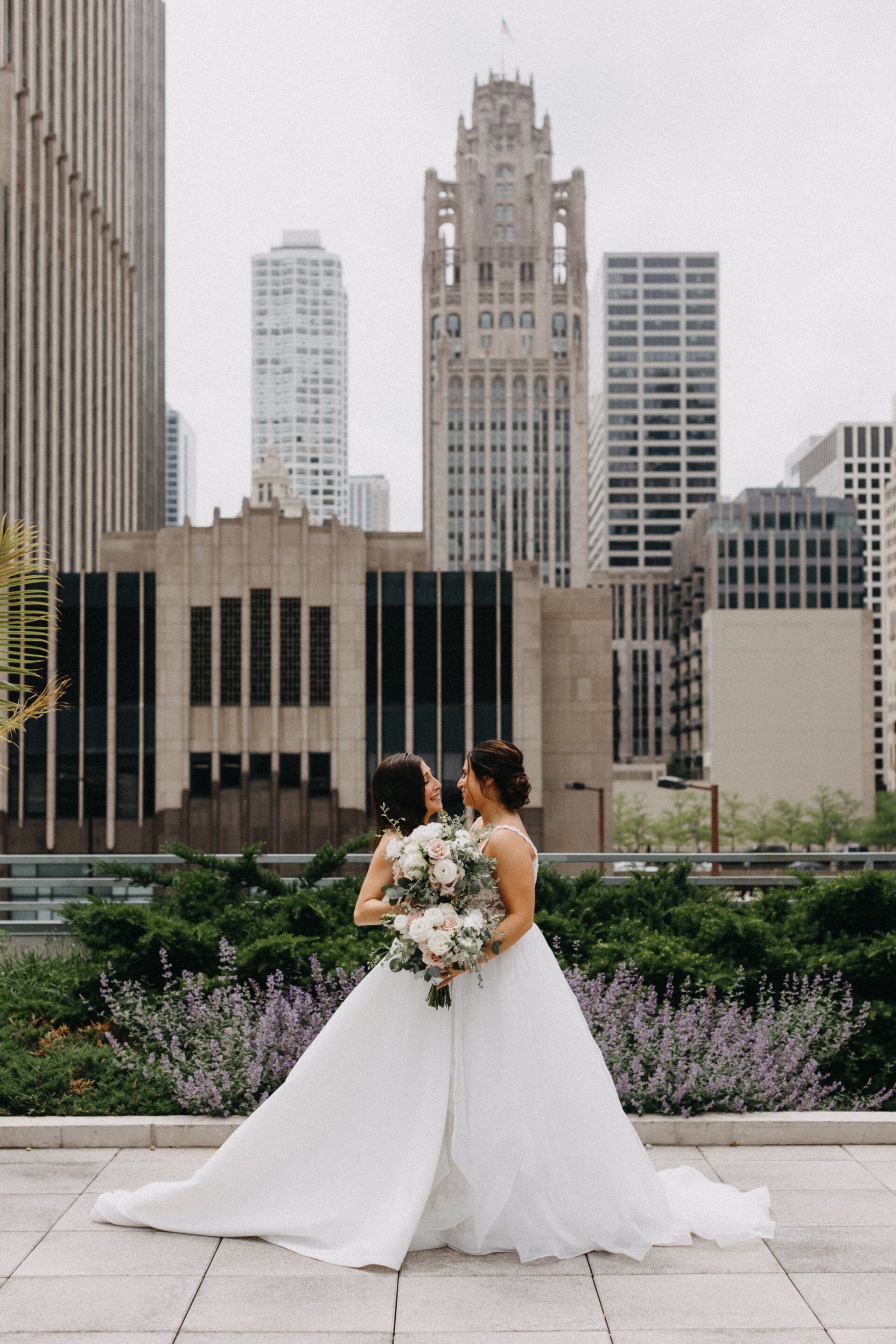 Two women in wedding dresses are standing next to each other in front of a city skyline.