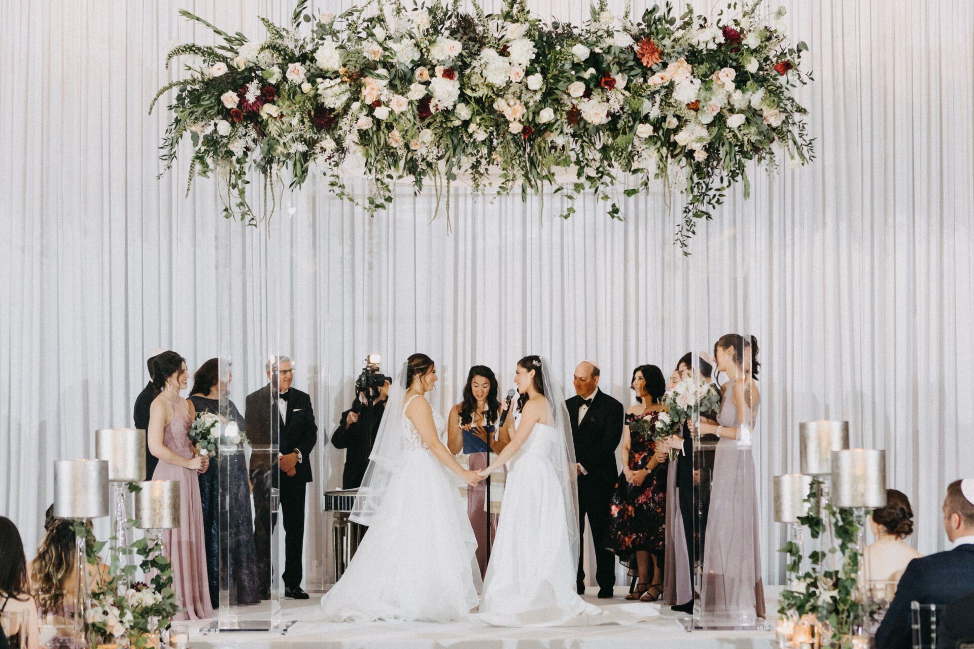 Two brides are holding hands at their wedding ceremony under a floral arch.
