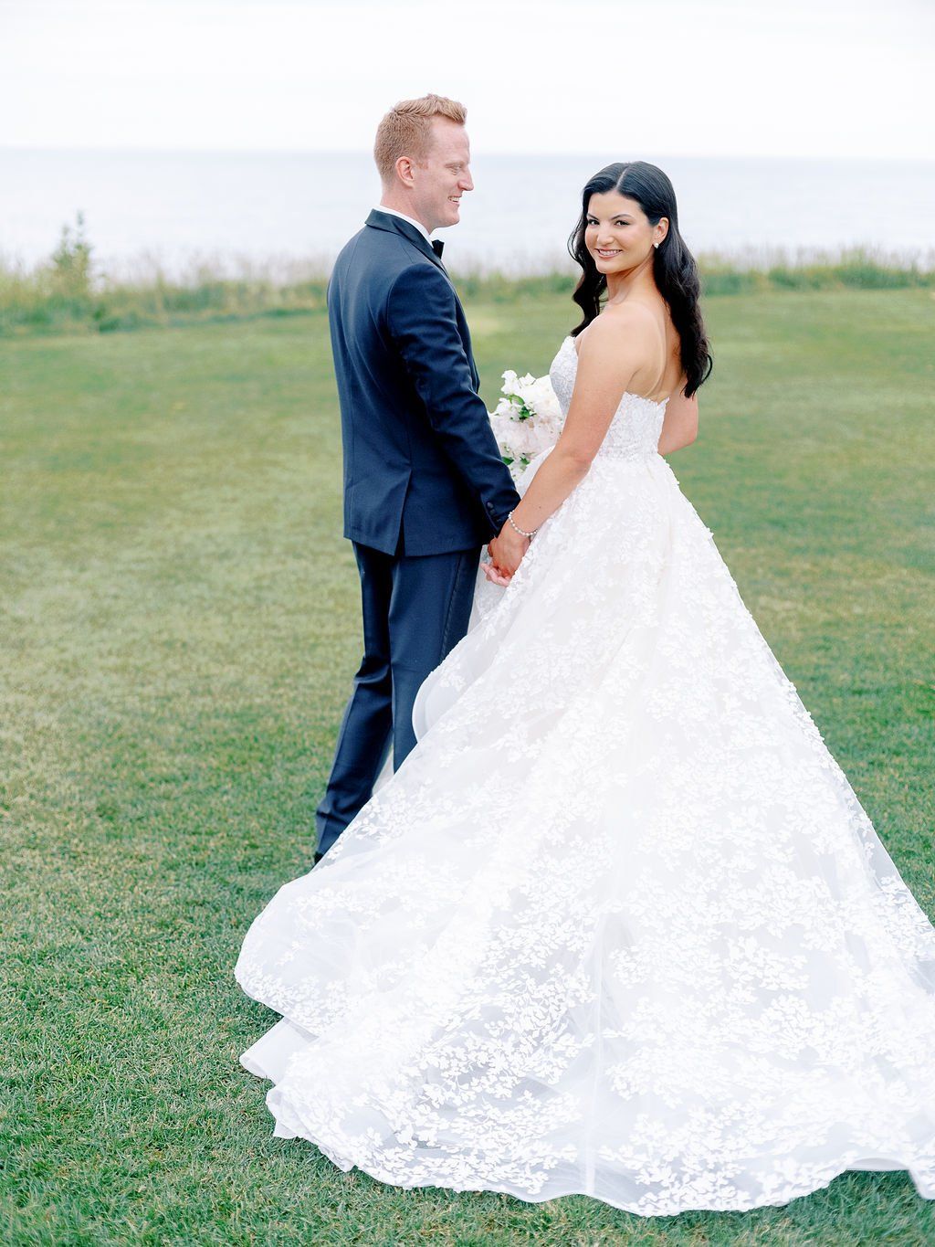 A bride and groom are standing in a grassy field holding hands.