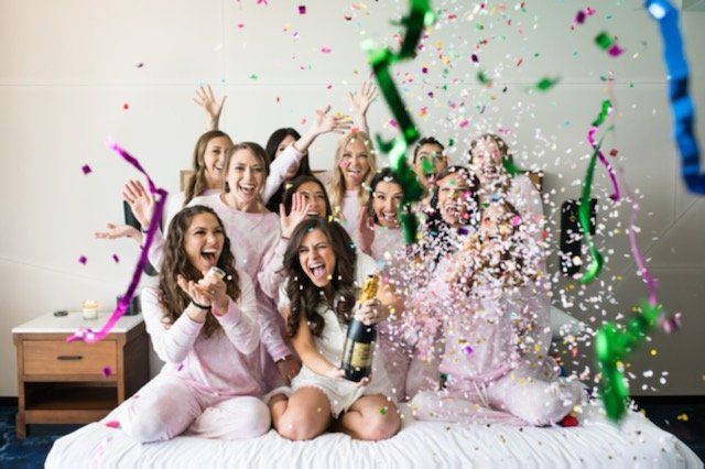 A group of women are sitting on a bed holding champagne bottles and throwing confetti.