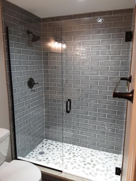 A walk-in shower with gray subway tile walls, a pebble floor, a glass door, and dark fixtures in a tiled bathroom.