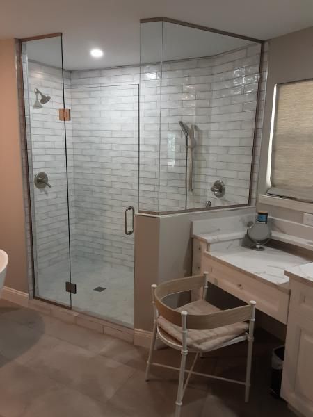 A glass-enclosed shower with white subway tiles, a showerhead, and a vanity area with a chair in a bathroom.
