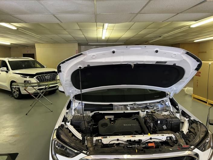 A white car with an open hood inside a workshop, with another white car parked in the background.