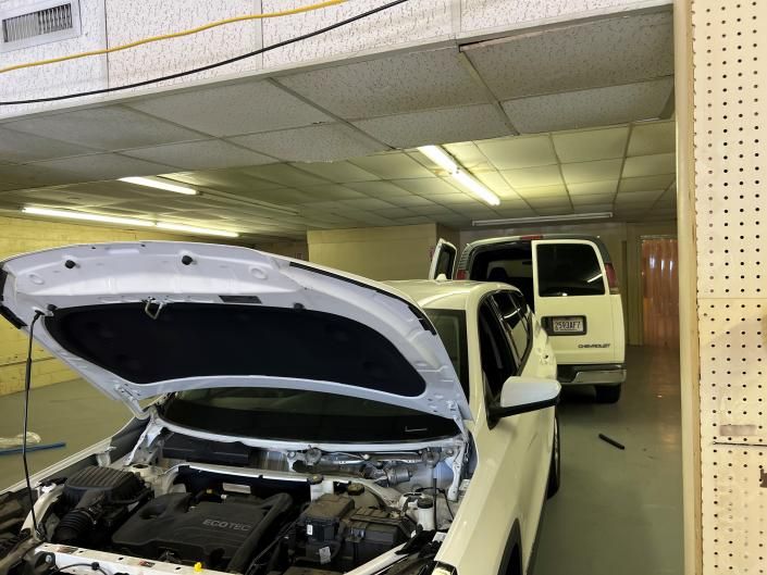 White car with its hood open in a repair shop, with a white van parked behind it.