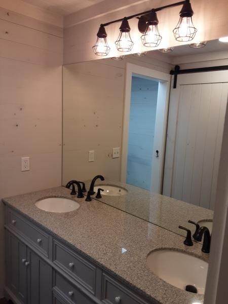 A modern bathroom vanity with grey cabinets, a speckled stone countertop, two sinks, and a large mirror with light fixtures.
