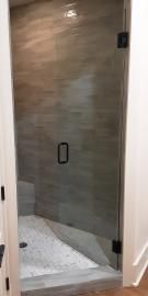 A glass shower door with a black handle and hinges, set against a tiled wall and a pebble-tiled shower floor.