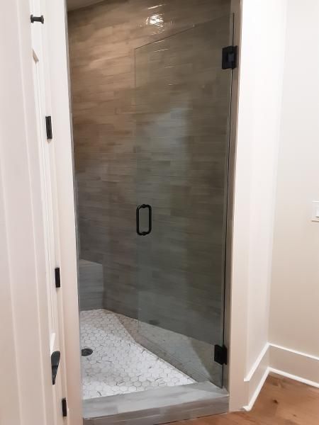 A glass shower door with a black handle and hinges, set against horizontal tan tile and a white pebble shower floor.