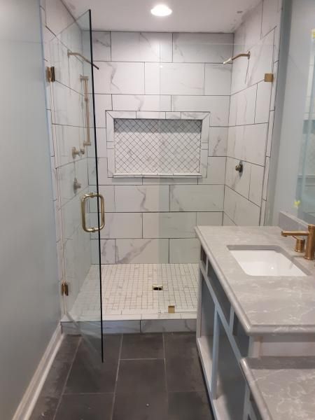 A walk-in shower with marble-look tiles, a glass door, a decorative mosaic niche, and a nearby grey vanity with gold faucet.