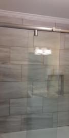 A glass shower door with a sliding metal track mounted against a wall of horizontal, light gray wood-look tiles.