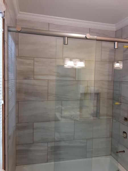 A sliding glass shower door mounted on a metal track, installed over a bathtub with gray, rectangular wall tiles.