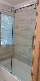 A frameless glass sliding shower door installed over a white bathtub with light beige, rectangular tiled walls.