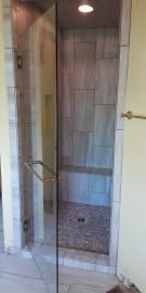 A walk-in shower with a glass door, light-colored tile walls, a built-in bench, and a pebbled floor.