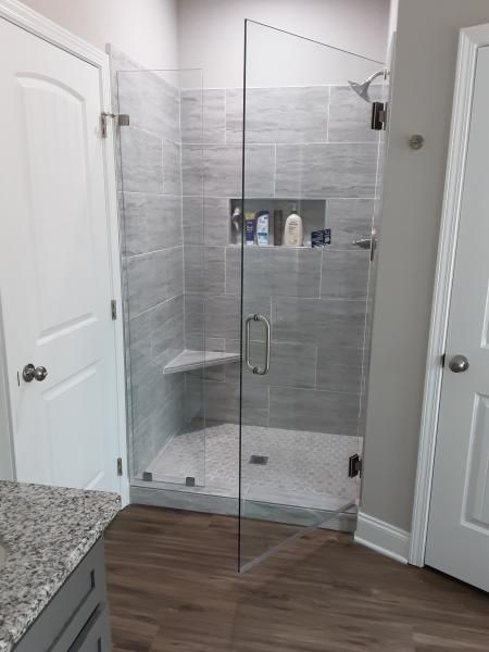 A glass-enclosed shower with grey tiled walls, a built-in corner bench, a recessed niche, and a light pebble-tiled floor.