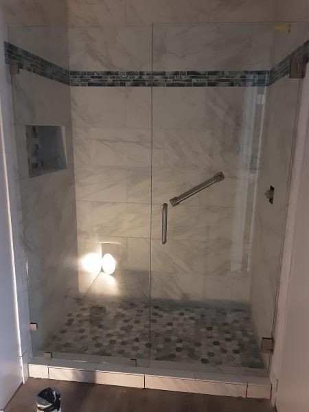 A walk-in shower with light marble-style wall tiles, a mosaic pebble floor, glass door, and a horizontal grab bar.