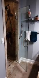 A glass shower door with a metal handle, positioned in a tiled bathroom with a towel rack and shelf nearby.