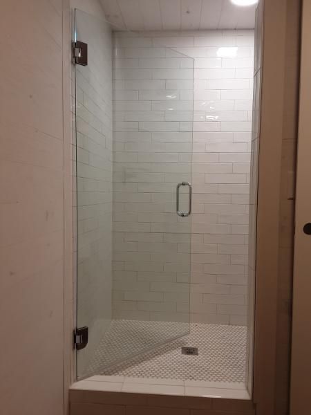 A frameless glass shower door with a metal handle and hinges installed in a white tiled bathroom.