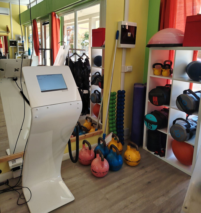 Sala fitness con kettlebell colorate, corde per esercizi e palle da ginnastica accanto a un chiosco con schermo tattile.