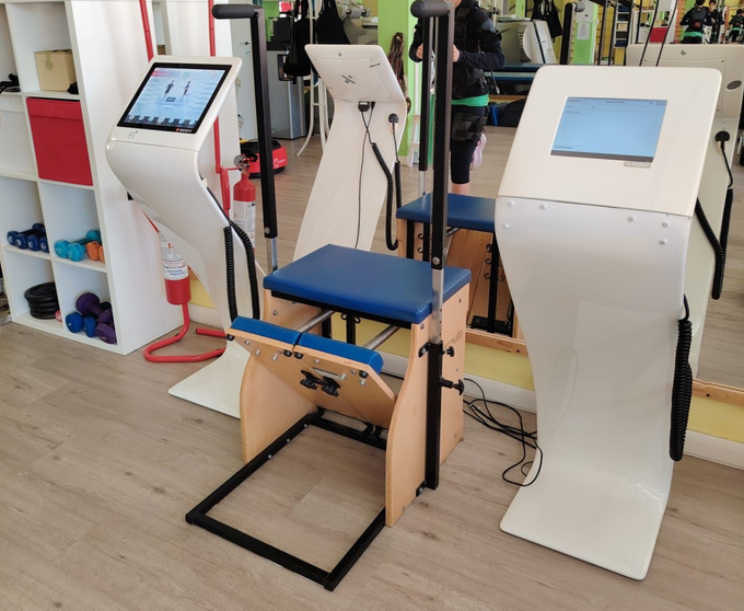Macchina per esercizi adattiva con sedile blu e touchscreen bianchi in una sala fitness o di riabilitazione.