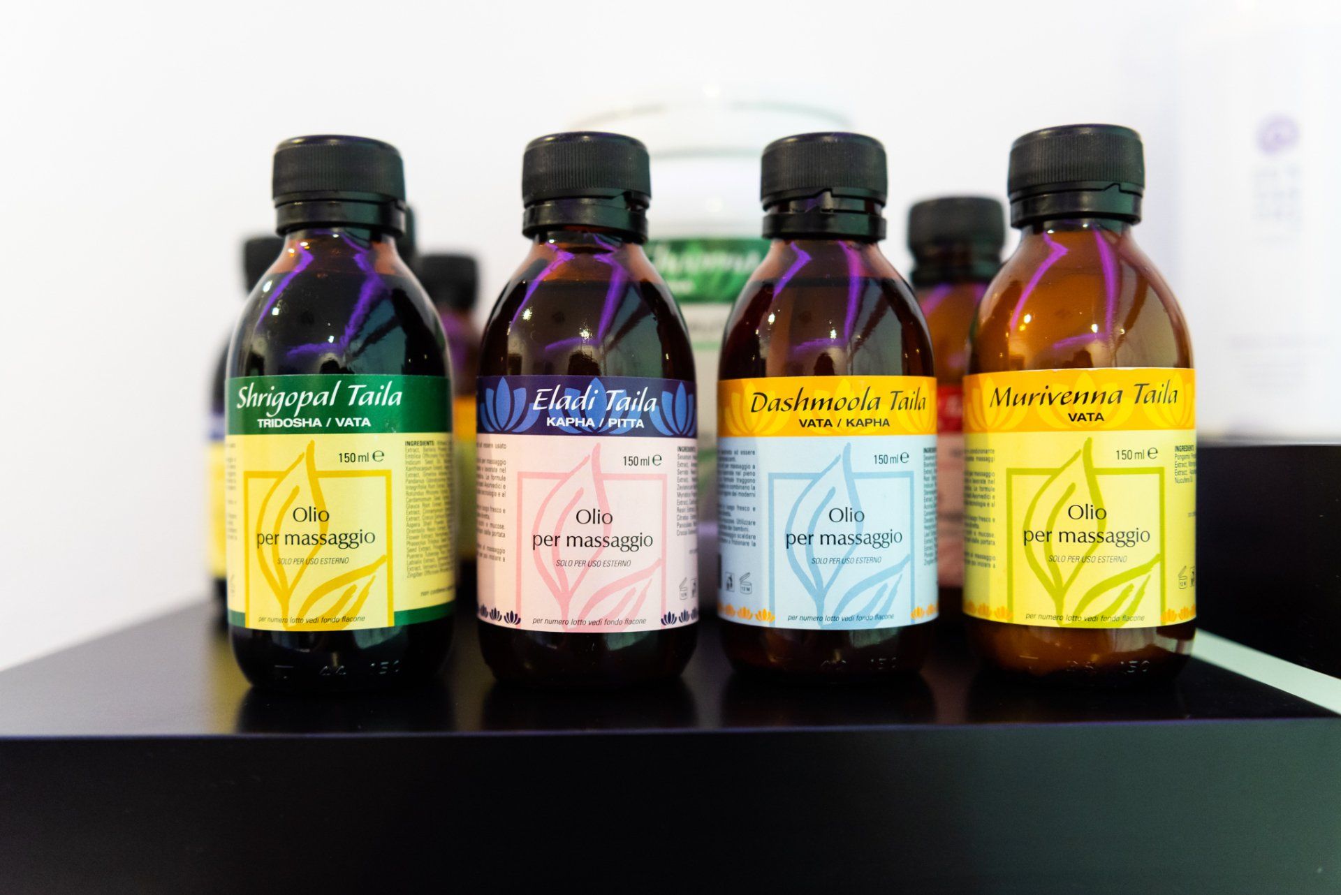 Su una superficie nera sono allineate quattro bottiglie marroni di olio da massaggio con etichette colorate.