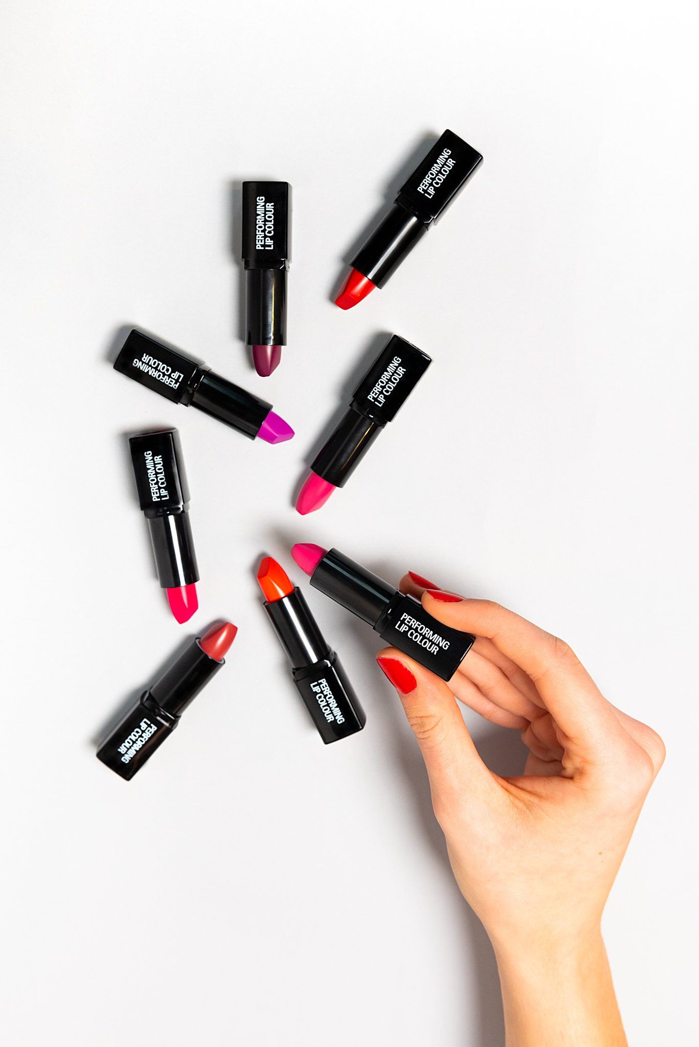 Una mano regge uno dei tanti tubetti di rossetto di vari colori sparsi su una superficie bianca.