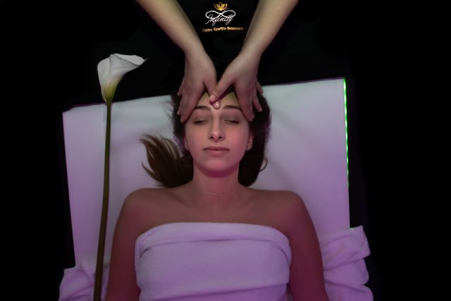 Una persona riceve un rilassante massaggio al viso mentre è sdraiata su un lettino bianco da spa, con una calla accanto.