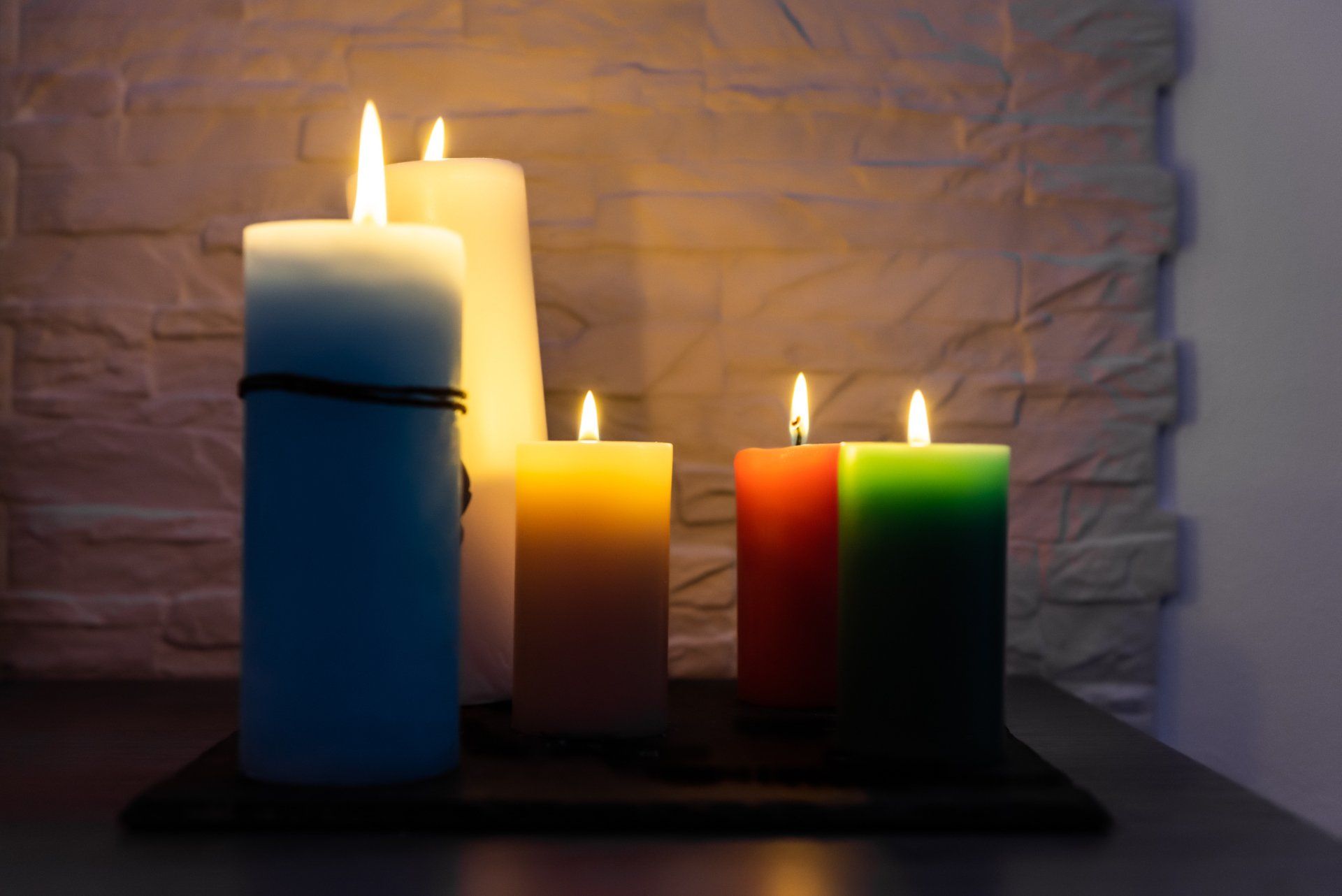 Cinque candele accese, una blu, una bianca, una gialla, una arancione e una verde, su una superficie scura contro un muro 