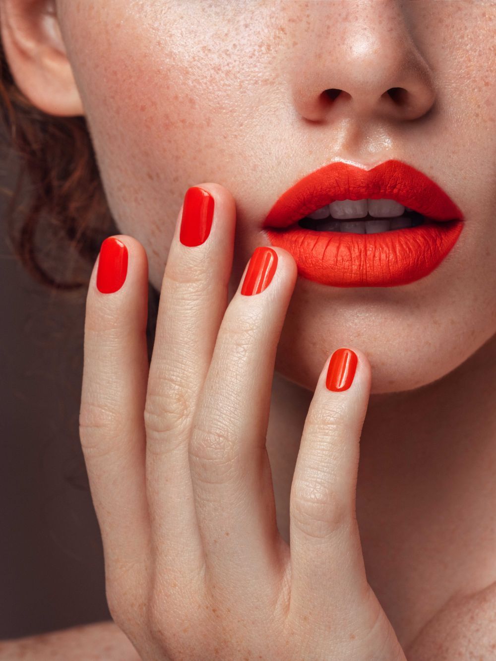 Un primo piano di una mano con unghie di un rosso acceso, vicino a un viso con un rossetto rosso brillante abbinato.
