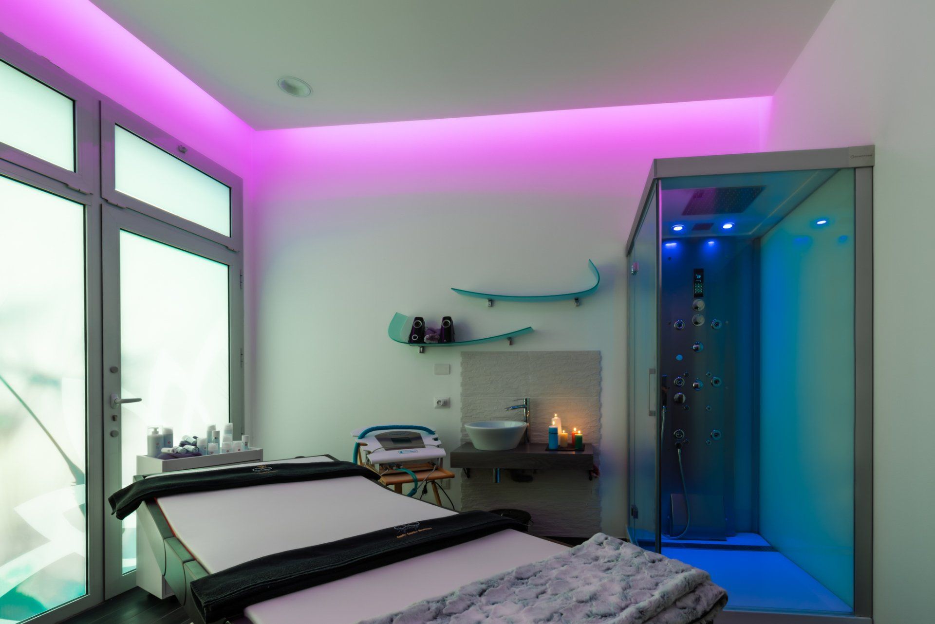 Una moderna sala spa con lettino da massaggio, doccia in vetro con illuminazione d'accento blu e illuminazione ambientale