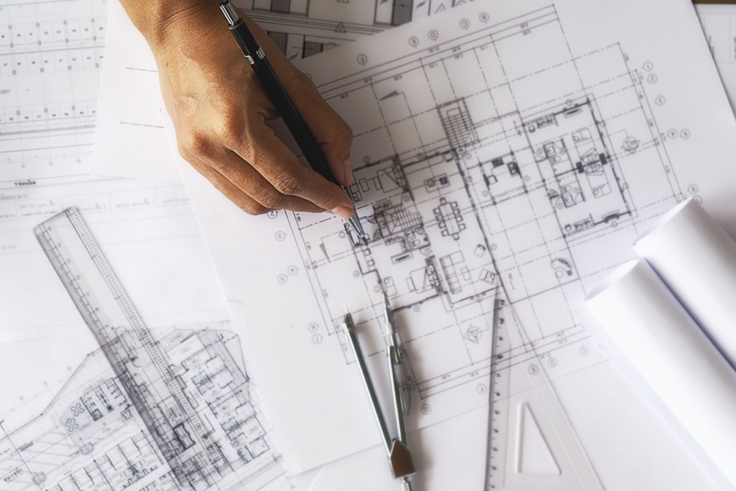 Top-down view of a hand using a pen to draw on architectural floor plans alongside drafting tools and rolled-up paper.
