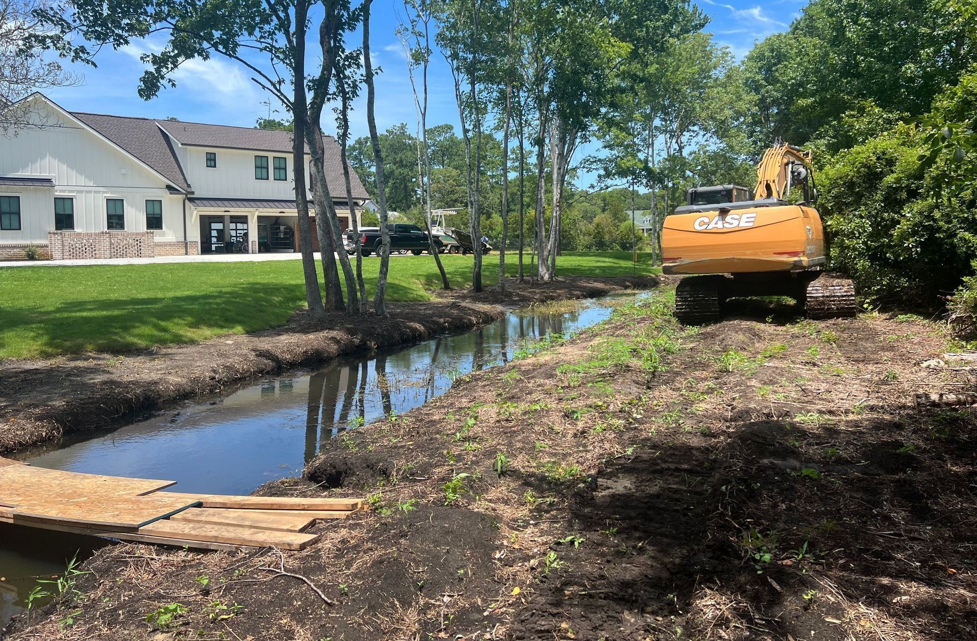 Excavator near a stream in a residential area, clearing the banks. A house with a green lawn is in the background.
