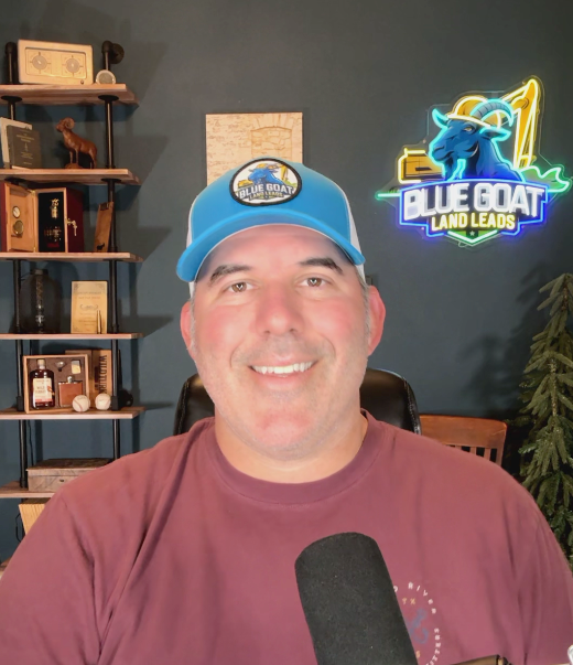 A man wearing a blue hat and a red shirt is holding a microphone in front of a blue goat land leads sign