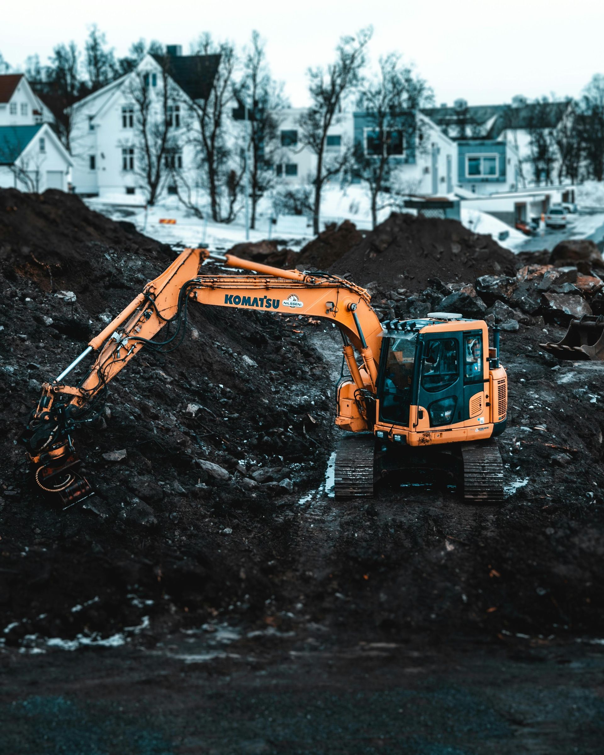 Yellow excavator on a dark pile of dirt, houses in the background.