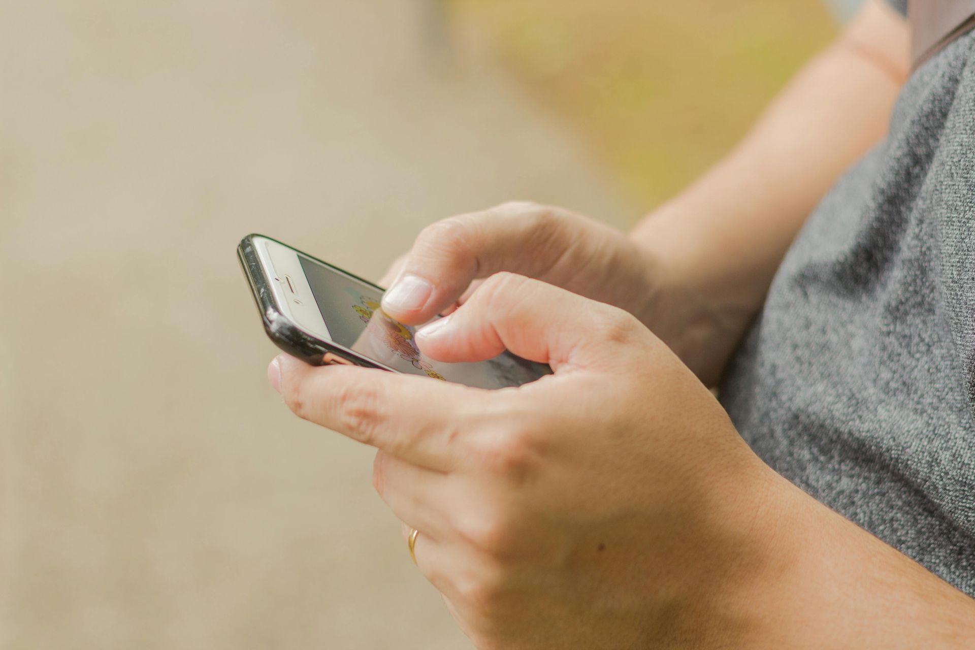 Person using a smartphone, typing with their thumbs. Grey shirt, blurry background.
