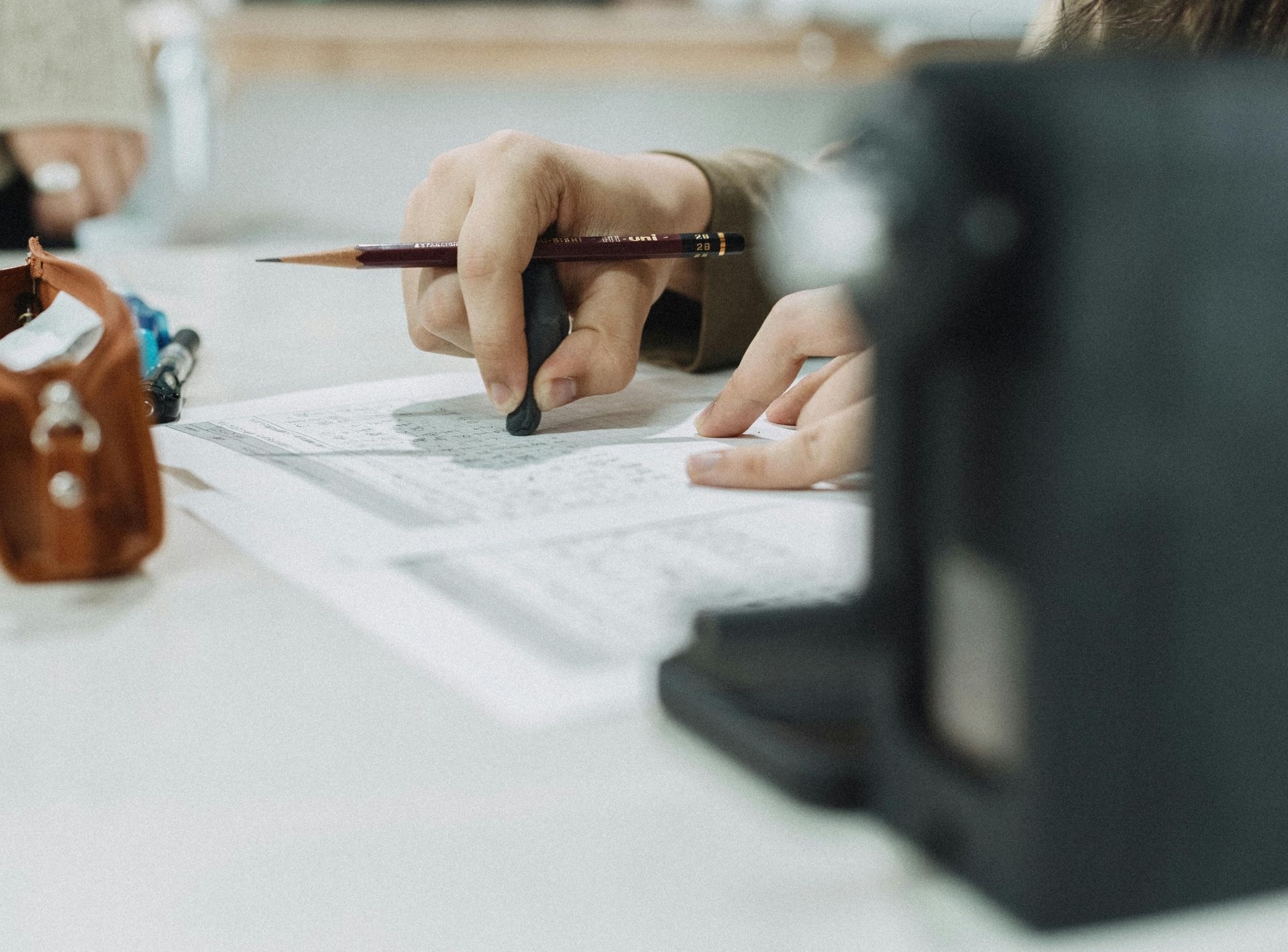 Person's hands sketching with a pencil and eraser on a white paper at a table. A camera is in the foreground.