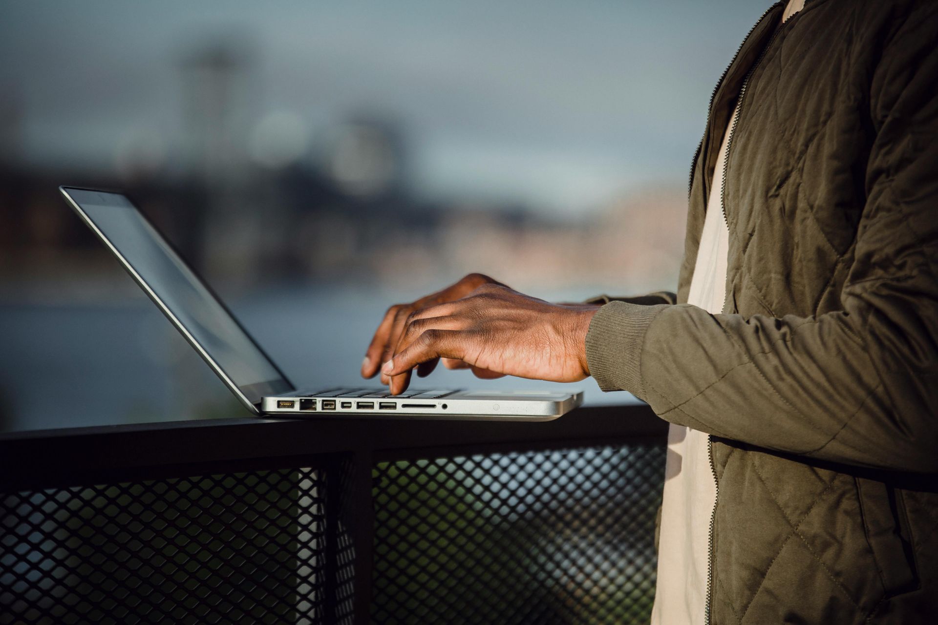 person typing on a laptop outdoors, wearing a green jacket, overlooking a blurred city and water.