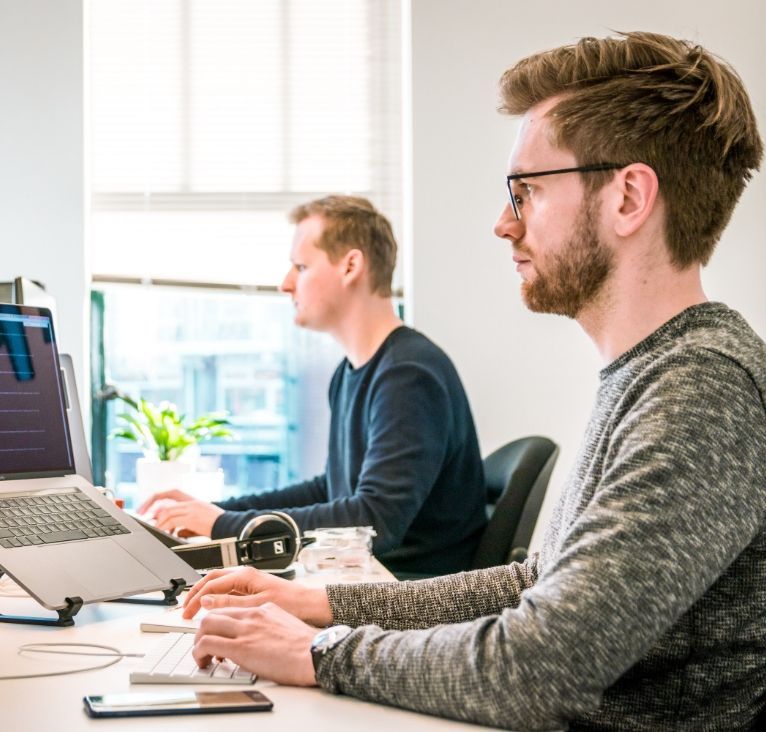 Two men working on laptops at a desk, one wearing glasses and typing, the other in the background.