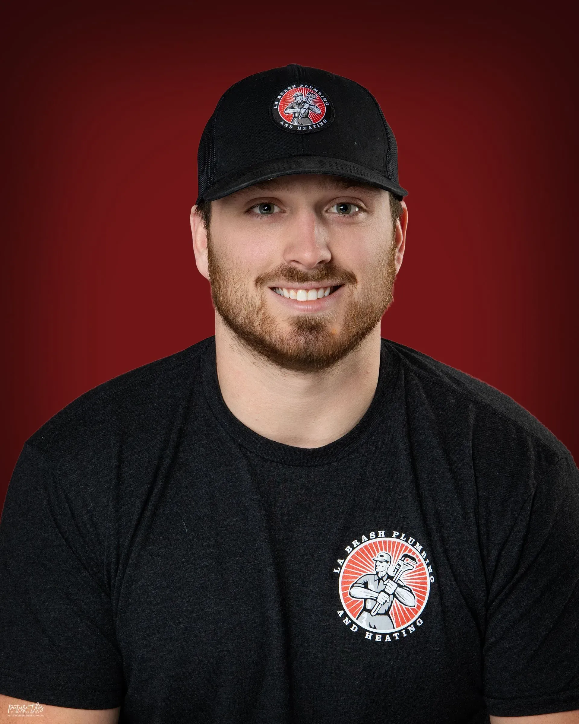 Man in black cap and T-shirt with a logo, smiling against a red background
