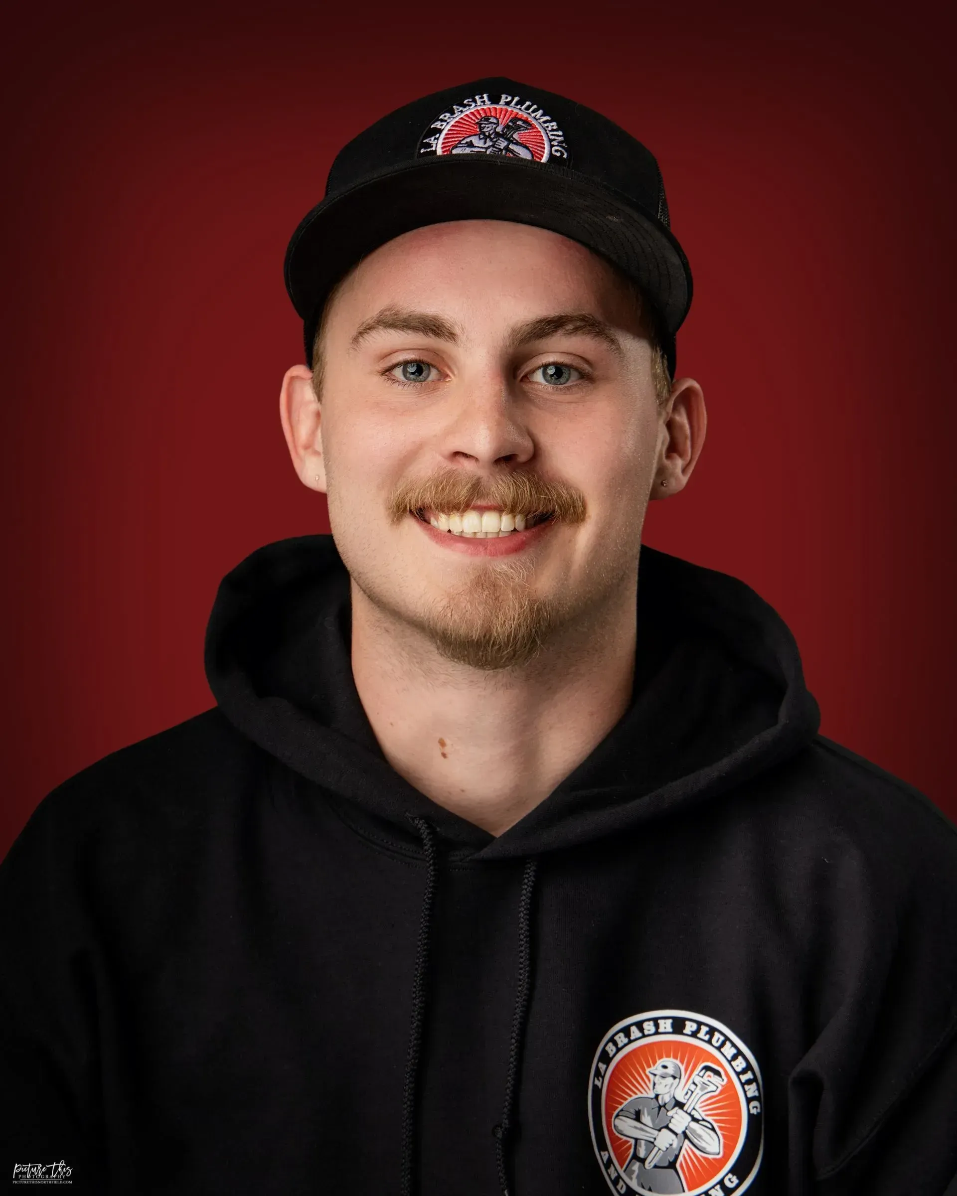 Smiling person in black hoodie and cap against a red background.