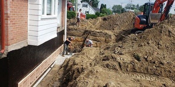 Des ouvriers effectuent des fouilles autour des fondations d'une maison à l'aide d'une excavatrice et de pelles.