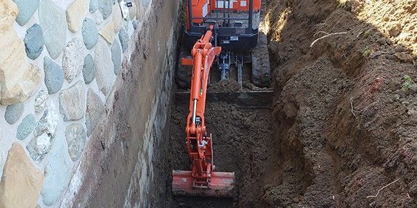 Une pelleteuse orange creuse une tranchée à côté d'un mur de pierre.