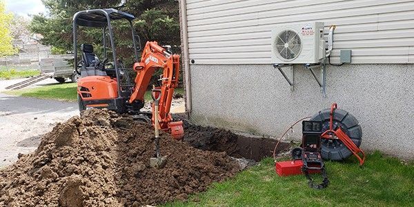 Une pelleteuse orange creuse une tranchée à côté d'un bâtiment. Unité de climatisation fixée au mur, matériel sur la pelouse.