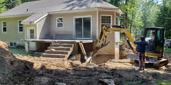 Une petite pelleteuse creuse près d'une maison, sous le regard d'un homme. Scène de chantier en extérieur.