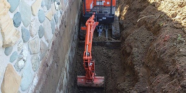 Une pelleteuse orange creuse une tranchée à côté d'un mur de pierre.