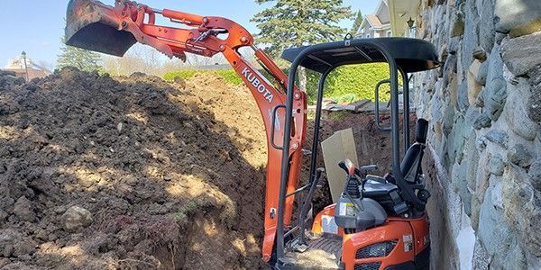 Une pelleteuse Kubota orange creuse une tranchée à côté d'un mur de pierre.