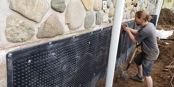 Un homme installe une membrane de drainage sur un mur en pierre. Il tient une pelle et travaille à l'extérieur.