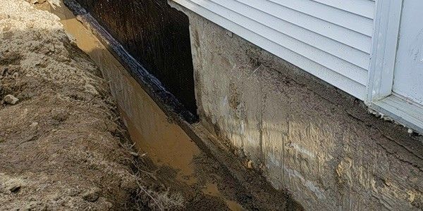 Une tranchée le long des fondations d'une maison, avec de la terre visible et une membrane noire.
